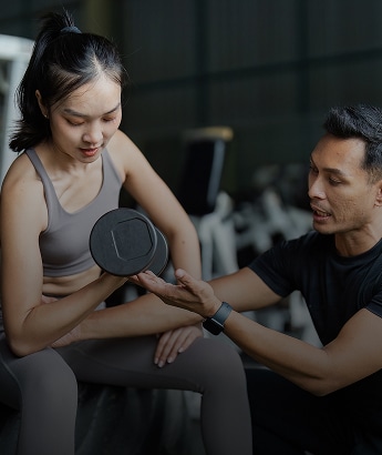 Femme asiatique s'entraînant avec un haltère au gym, coach sportif masculin expliquant le mouvement. Elle est concentrée, il la guide attentivement.