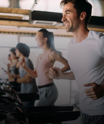 Hommes et femmes souriants courent sur des tapis de course en salle de sport. Un homme joyeux est au premier plan.