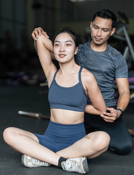 Femme étirant son bras assistée par un coach sportif dans une salle de gym. Elle est assise en tenue de sport, il guide son mouvement.